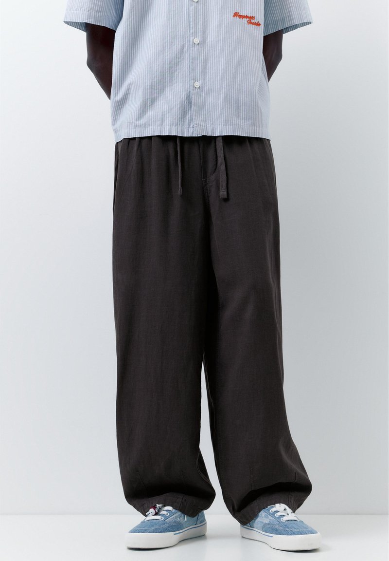 Personne portant un pantalon noir ample, une chemise à manches courtes rayée bleu clair et des baskets bleu clair sur un fond blanc uni.