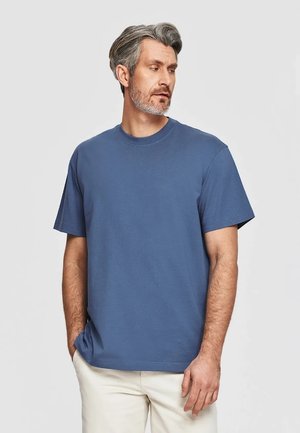 Mężczyzna z siwymi włosami i brodą, ubrany w niebieski t-shirt z krótkim rękawem i jasne spodnie, stoi z jedną ręką w kieszeni.