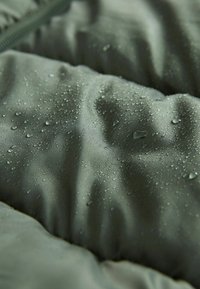 Vandeniui atsparus audinys muted žalios spalvos, turintis tekstūruotą paviršių su matomais lašeliais, kurie rodo drėgmei atsparumą ir dinamiškas dvejopes formas.