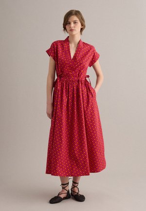 Mujer de pie con vestido midi rojo con patrón geométrico naranja, mangas cortas, escote cruzado y lazos laterales, combinado con zapatos planos negros con cordones.