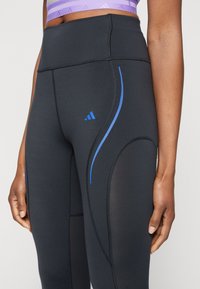 Leggings noirs taille haute avec des accents courbés bleu, fabriqués en tissu extensible. Présentent des coutures plates et un logo brodé.
