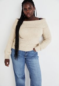 Sieviete ar gariem bizēm valkā krēmkrāsas plecus atklātu džemperi un zilas džinsas, stāvot ar vienu roku kabatā pret baltu fonu.