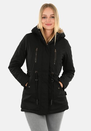 Junge Frau trägt eine schwarze Winterjacke mit Kapuze und Pelzbesatz, Hände in den Taschen, steht vor einem weißen Hintergrund.