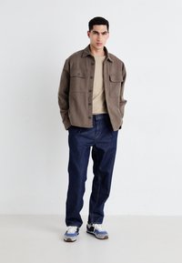 Mężczyzna stojący z rękami w kieszeniach, noszący brązową zapinaną kurtkę na beżową koszulę, ciemnoniebieskie dżinsy i niebiesko-białe trampki.