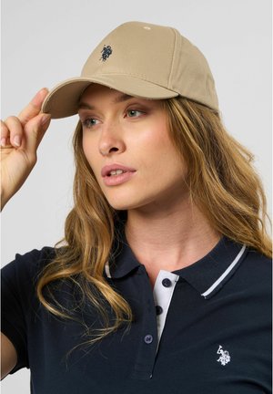 Khaki baseball cap with stitched logo, curved brim, and adjustable back. Worn with a navy polo featuring white accents and buttons.