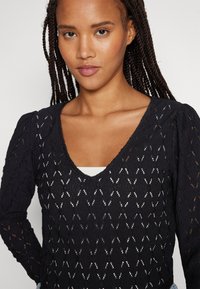 Μαύρο πλεκτό τοπ με V λαιμό, που διαθέτει υφή και φουσκωμένα μανίκια. Το ύφασμα παρουσιάζει μια λεπτεπίλεπτη δαντελένια σχεδίαση με ανοιχτές λεπτομέρειες.