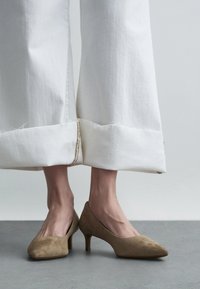 Sapatos de salto alto em camurça castanho claro com bicos arredondados, combinados com calças brancas de perna larga com bainhas desfiadas e costura decorativa.