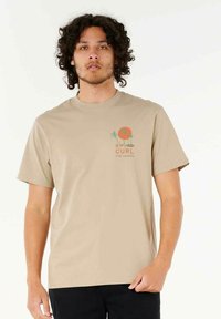 Camiseta de algodón beige de manga corta y cuello redondo. Presenta un pequeño gráfico de un atardecer, palmeras y texto en el lado izquierdo del pecho.