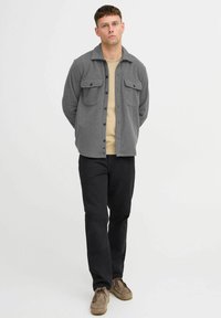 Gray button-up shacket with two chest pockets, worn over a beige top. Paired with black pants and light brown shoes. Minimalistic design.