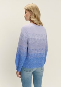 Kobieta z blond włosami, ubrana w sweter o gradientowej fakturze w odcieniach fioletu i niebieskiego, połączony z jasnoniebieskimi dżinsami, zwrócona bokiem.