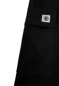 Schwarzes Stoffkleidungsstück mit einer quadratischen Tasche. Auf der Vorderseite befindet sich ein weißes Etikett mit einem kreisförmigen Logo und dem Text "HONEST RULES". Glatte Textur, taillierte Passform.