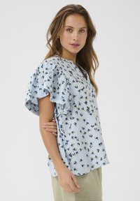 Svetlomodrá kvietkovaná blúzka s volánikmi na krátkych rukávoch, s malými zelenými a námorníckymi kvetmi, V-krkom a mäkkou, štruktúrovanou tkaninou.