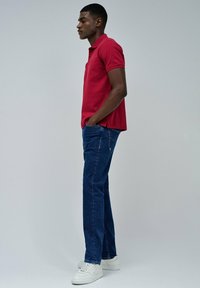 Maglietta polo rossa a maniche corte, abbinata a jeans blu scuro e sneaker bianche. L'outfit presenta una vestibilità casual e una texture liscia.