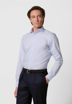 Hombre con camisa azul claro abotonada, pantalones oscuros y cinturón negro, de pie con una mano en el bolsillo, mirando hacia un lado.