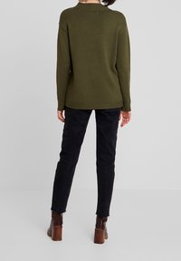 Olivgrüner Strickpullover mit entspannter Passform, geripptem Kragen und Bündchen, kombiniert mit schwarzen schmal geschnittenen Hosen und braunen Stiefeln mit Absatz.