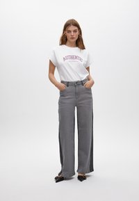 Witte T-shirt met de tekst "AUTHENTIC", gecombineerd met hooggerekende, wijde grijze jeans met zijkantenaccenten en zwarte puntige schoenen.
