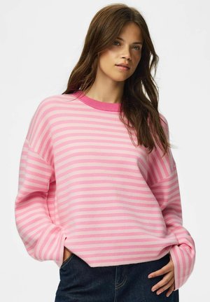 Jeune femme aux longs cheveux bruns portant un pull rayé rose et rose clair et un jean bleu foncé, posant avec une main dans la poche.