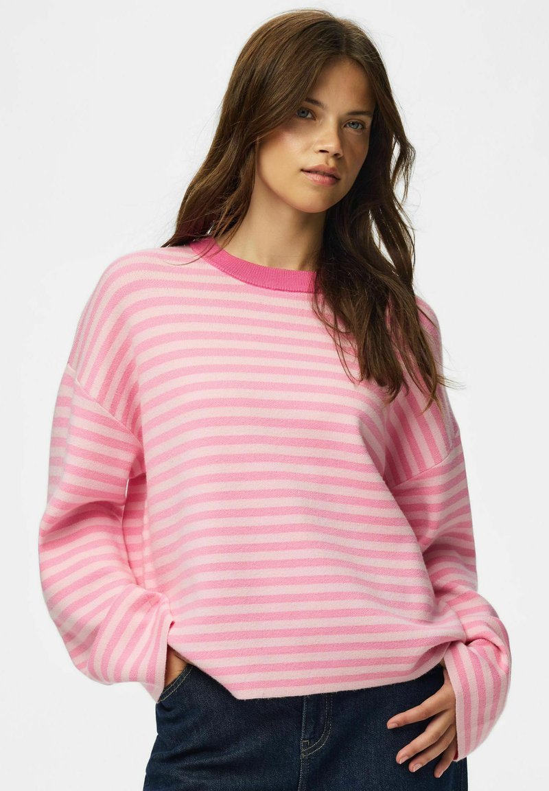 Jeune femme aux longs cheveux bruns portant un pull rayé rose et rose clair et un jean bleu foncé, posant avec une main dans la poche.