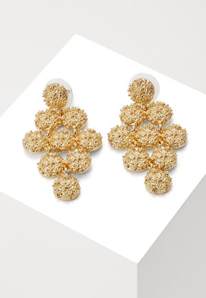 Złote kolczyki w kształcie okrągłych klastrów o fakturze, ułożone w wielowarstwowy geometryczny wzór na białej powierzchni.