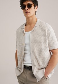 Camicetta corta beige chiaro con maniche corte e pattern geometrico testurizzato, bottoni frontali e colletto rilassato, abbinata a una canotta bianca sottostante.
