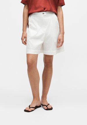 Person iført hvide knælange skræddersyede shorts, en rustfarvet kortærmet skjorte og sorte flip-flops, stående mod en hvid baggrund.