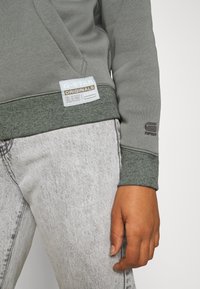 Zielona bluza z ribbowanymi mankietami, z naszywką logo na dolnym brzegu. Noszona z jasnoszarymi dżinsami, pokazującymi szczegóły tekstury tkaniny.