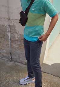 Person som bär en turkosmönstrad skjorta, mörka jeans, vita sneakers och en svart axelremsväska står på en betongyta nära en vägg.