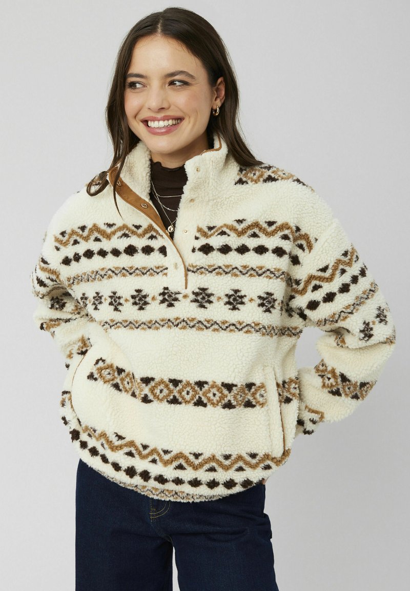 Pull en polaire crème avec des motifs géométriques marron et beige. Présente une fermeture éclair mi-hauteur, un col et une poche kangourou. Tissu doux et texturé.