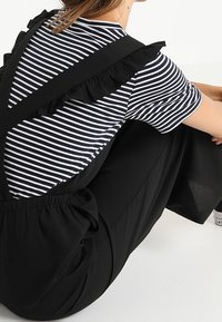 Czarne ogrodniczki mają falbany na ramiączkach, noszone na granatowej i białej w paski koszulce. Strój ma luźny krój i miękką teksturę.