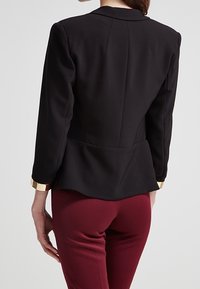 Blazer noir avec épaules structurées, design peplum et accents dorés sur les poignets. Porté sur un pantalon bordeaux ajusté. Texture de tissu lisse.