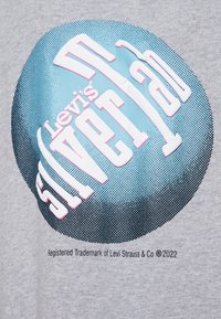 Camiseta de tela gris con un gráfico redondo azul que presenta "Levi's SilverTab" en texto blanco y rosa, junto con detalles de marca en la parte inferior.