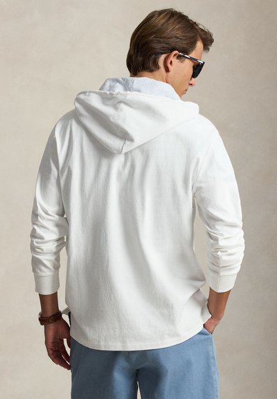 Homme portant un sweat à capuche blanc, un short bleu, des lunettes de soleil et une montre-bracelet, debout les mains dans les poches, dos à nous.