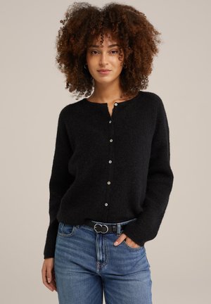 Cardigan noir en tricot texturé avec un col rond et un bouton. Porté sur un jean bleu, il présente une fine ceinture avec une boucle circulaire.