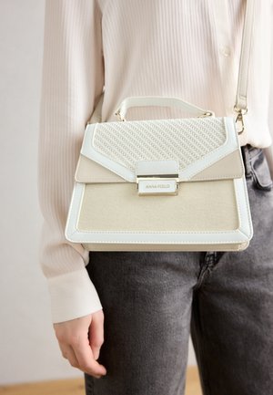 Kvinde iført en lysebeige textured håndtaske med tophåndtag og guldlås, kombineret med en cremefarvet bluse og mørkegrå jeans.