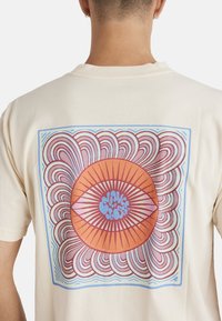 Camiseta color crema con un gran gráfico circular con patrones en naranja y rosa, rodeado de líneas onduladas y acentos en azul.