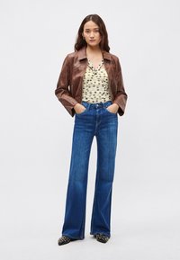 Barna bőrkabát krémszínű, pöttyös felső fölött, hozzá bővülő kék farmernadrág és fekete, ezüst díszítésű hegyes orrú cipők.