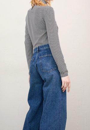 Persona con una blusa de manga larga a cuadros en blanco y negro y jeans azules de tiro alto y pierna ancha, de pie de lado contra un fondo sencillo.