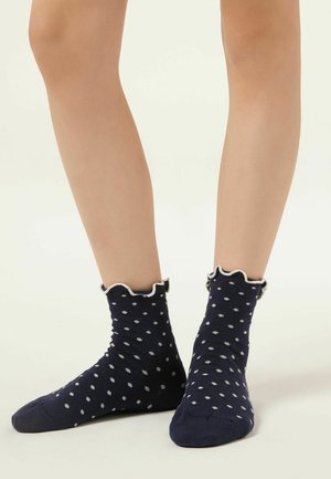 Pár nohou obutých v tmavě modrých kotníkových ponožkách s bílými puntíky a volánkovým bílým lemem nahoře na jednobarevném pozadí.