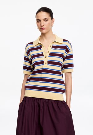 Femme portant un polo jaune à manches courtes avec des rayures horizontales marron, bleu et blanc et une jupe violet foncé, les mains dans les poches.