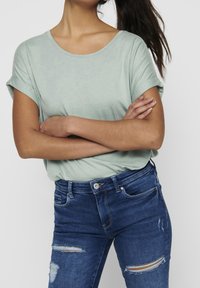 Jasnozielony t-shirt z krótkim rękawem i zaokrąglonym dekoltem. Połączony z dopasowanymi niebieskimi dżinsami z przetarciami i zapinanymi na guziki z przodu.