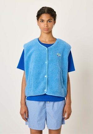 Blue fleece vest with a rounded neckline, front buttons, and side pockets. Worn over a blue shirt and paired with striped shorts.