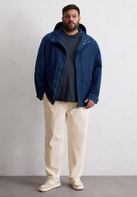 Marineblaue leichte Jacke mit Kapuze, zwei Fronttaschen, kombiniert mit einem dunklen T-Shirt, crèmefarbener Hose und beigen Sneakers.