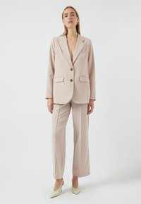 Vaalea beige puku takki, jossa on viistetyt kulmat, kaksi nappia ja taskut, yhdistettynä samanvärisiin räätälöityihin housuihin. Sujuva kangas, rento istuvuus.