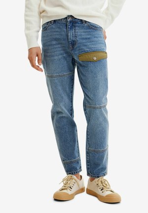 Personne portant un jean en denim bleu avec une poche boutonnée verte sur la cuisse et des baskets beiges avec des semelles en gomme.