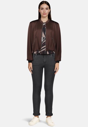 Brązowa kurtka bomberka z satynowym wykończeniem, zapinana na zamek, z kieszeniami na suwak, ściągaczami na rękawach, zestawiona z wzorzystą bluzką i ciemnymi dżinsami, wykończona białymi trampkami.