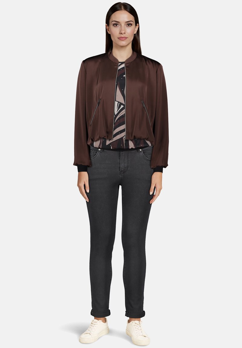 Brun bomberjakke med satinfinish, lynlåslommer, ribbede manchetter, parret med en mønstret top og mørke jeans, afsluttet med hvide sneakers.