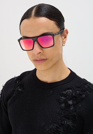 Giovane con capelli lunghi e scuri legati indietro, indossa occhiali da sole riflettenti rosa, maglione nero lavorato a maglia consumato e un piccolo orecchino a cerchio.