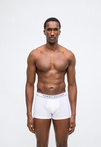 Αθλητικός άνδρας στέκεται, φορώντας λευκά μπόξερ Tommy Hilfiger σε απλό ανοιχτό φόντο.