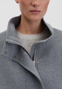 Nahaufnahme einer Person, die einen grauen Wollmantel in Schichten mit steigendem Kragen trägt, der teilweise offen ist, über einem hellgrauen Pullover.