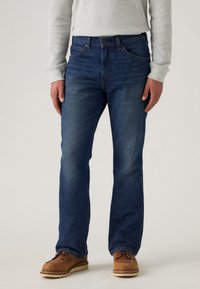 Jeans de mezclilla azul oscuro con un diseño de pierna recta, costuras prominentes y un estilo clásico de cinco bolsillos. Llevados con zapatos de cuero marrón.